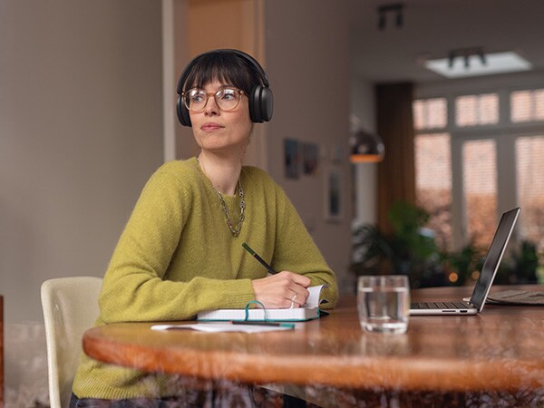Persona, kas valkā ērtas Philips austiņas, sēž pie galda ar piezīmjdatoru un portatīvo datoru