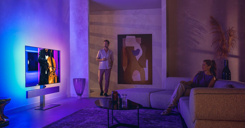 Vīrietis un sieviete stilīgā viesistabā skatās Philips Ambilight televizoru, kurā tiek rādīts čella priekšnesums