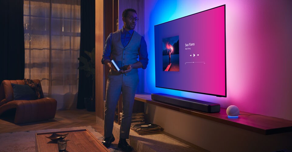 Cilvēks modernā viesistabā blakus Philips Ambilight televizoram ar mūzikas saskarni un Fidelio skaņas joslu