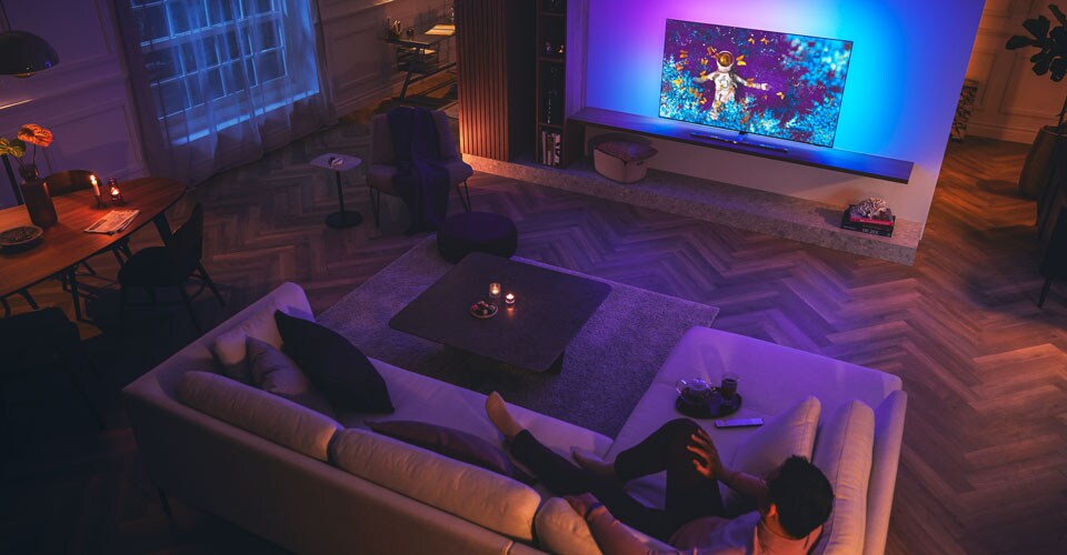 Vīrietis atpūšas uz dīvāna vāji apgaismotā viesistabā, skatoties Philips Ambilight televizoru