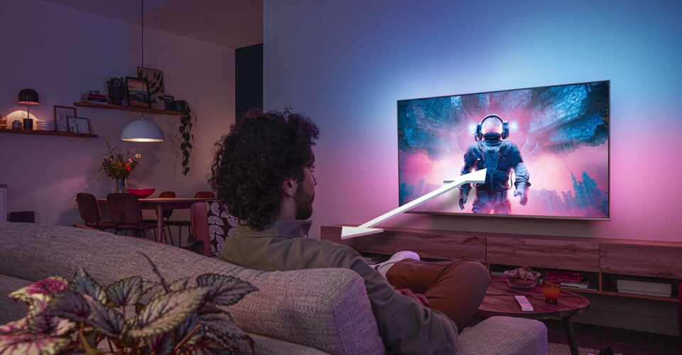 Vīrietis sēž uz dīvāna un skatās Philips Ambilight televizoru, kurā redzama krāsaina astronauta aina, ar bultiņu, kas norāda optimālo skatīšanās attālumu