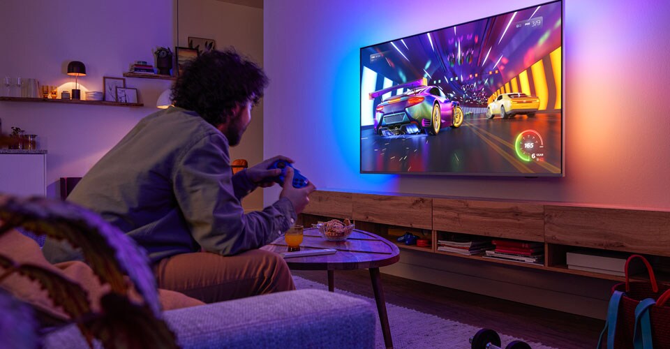 Cilvēks sēž uz dīvāna un spēlē sacīkšu videospēli Philips Ambilight televizorā
